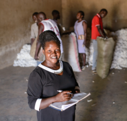 Ugandan woman stands at the centre of the photo smiling up at the camera as she takes notes. Blurred behind her are men packing cotton.