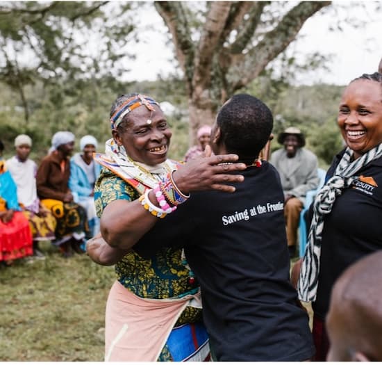 Two women hugging, one wearing Savings at the Frontier t-shirt, others smiling nearby.